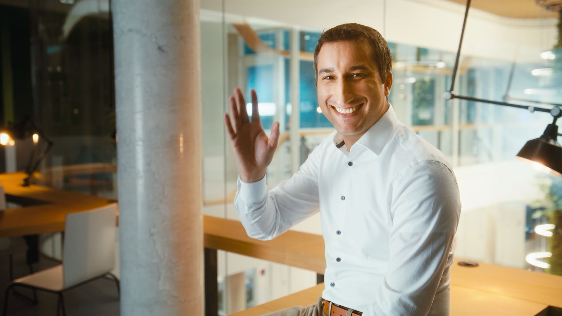 Lächelnder Mann in heller Business-Kleidung winkt in einer modernen Büroumgebung.
